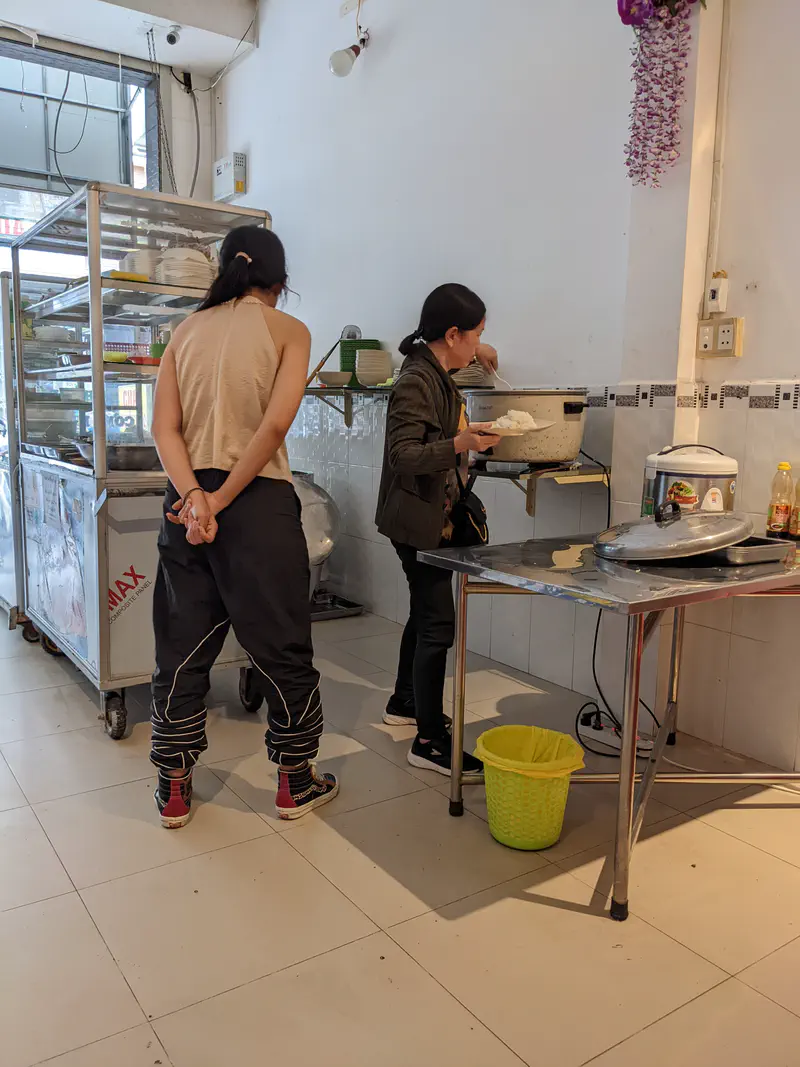 Woman serving rice from a large rice cooker inside a simple restaurant while another customer waits.