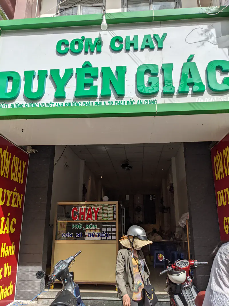 Front of a vegetarian restaurant named Cơm Chay Duyên Giác in Châu Đốc, An Giang, with a person wearing a sun hat and helmet standing outside near a motorbike.