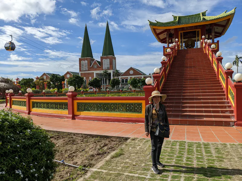 Woman wearing a sun hat standing in front of a temple staircase with traditional Asian architecture, with a church and a cable car in the background.