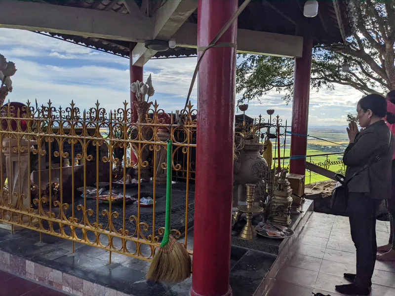 Woman praying at an outdoor altar with incense and offerings, overlooking rice fields.