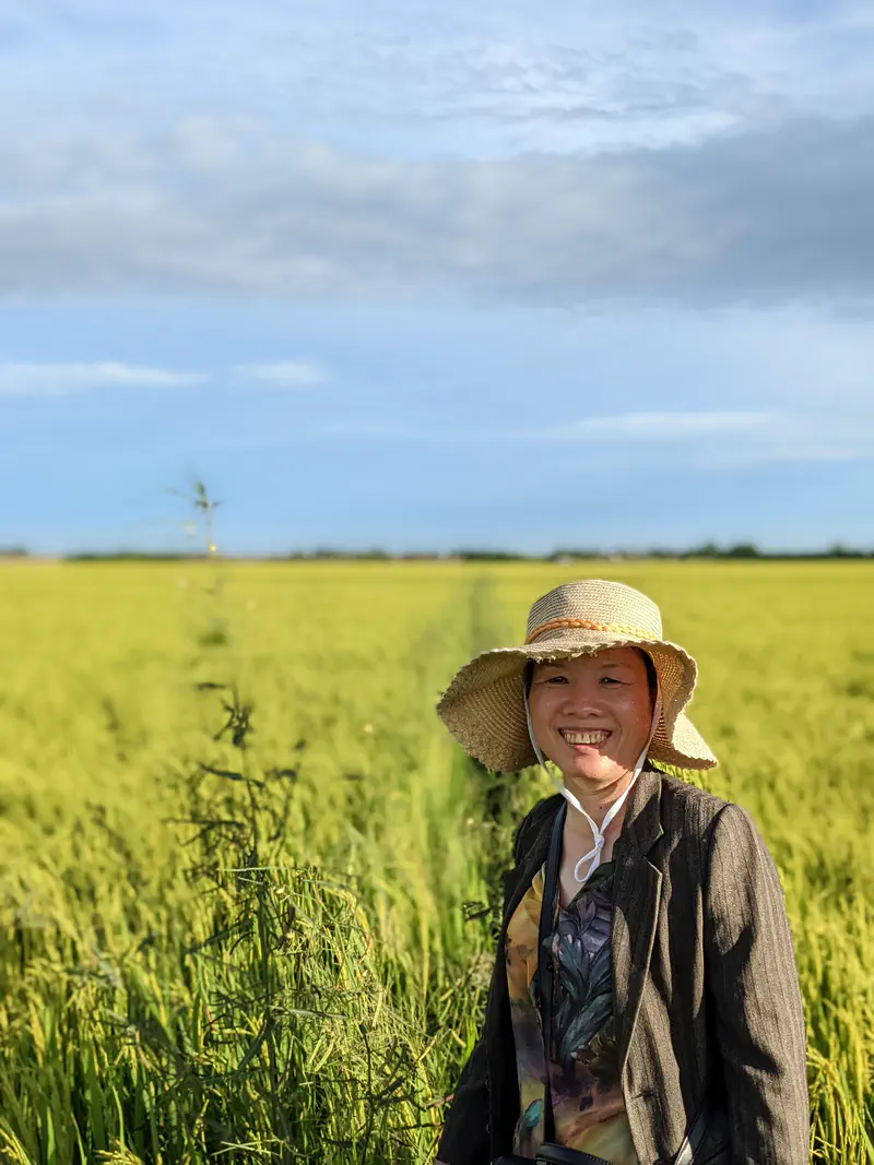 Smiling woman wearing a straw hat standing in a rice field under a partly cloudy sky.