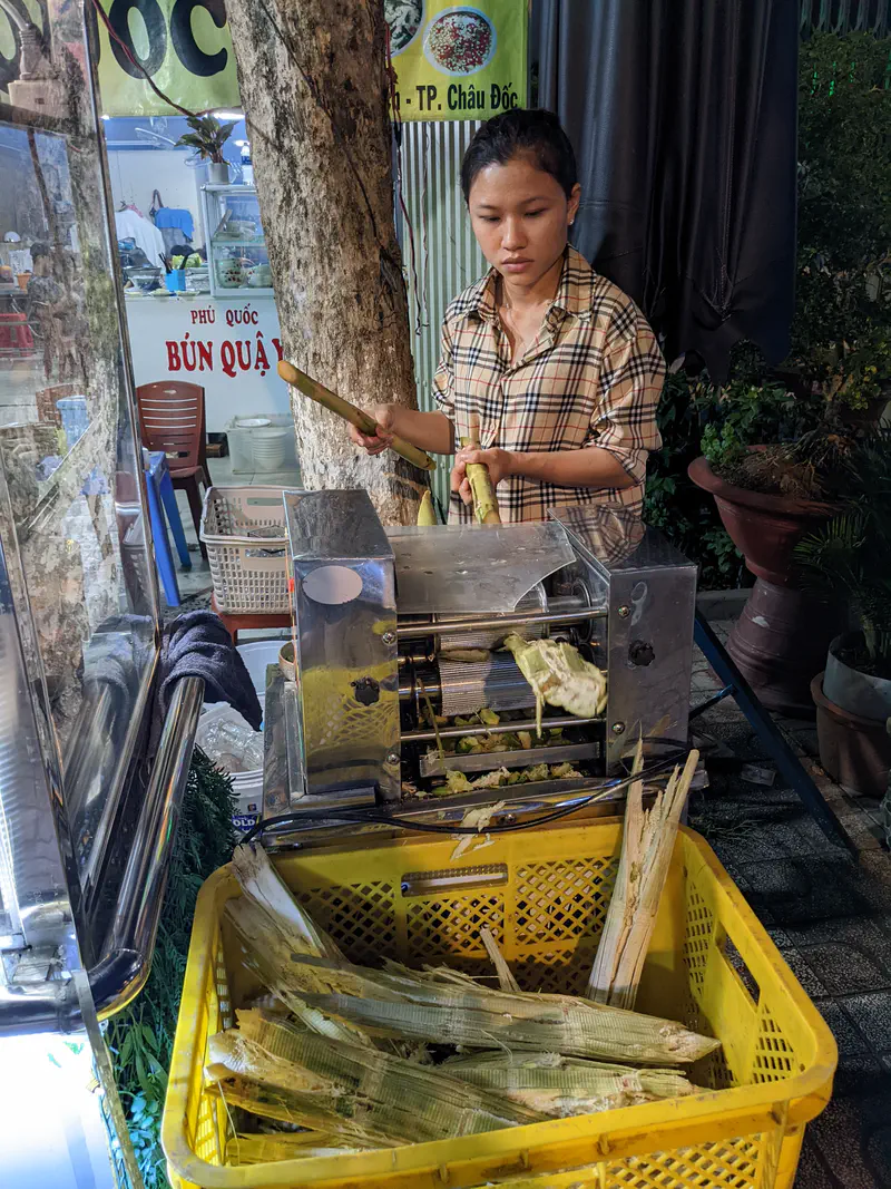 Woman operating a machine to press sugarcane for fresh juice at a street stall.
