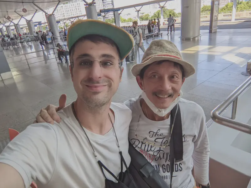Two men smiling for a selfie at an outdoor terminal with people and luggage carts in the background.