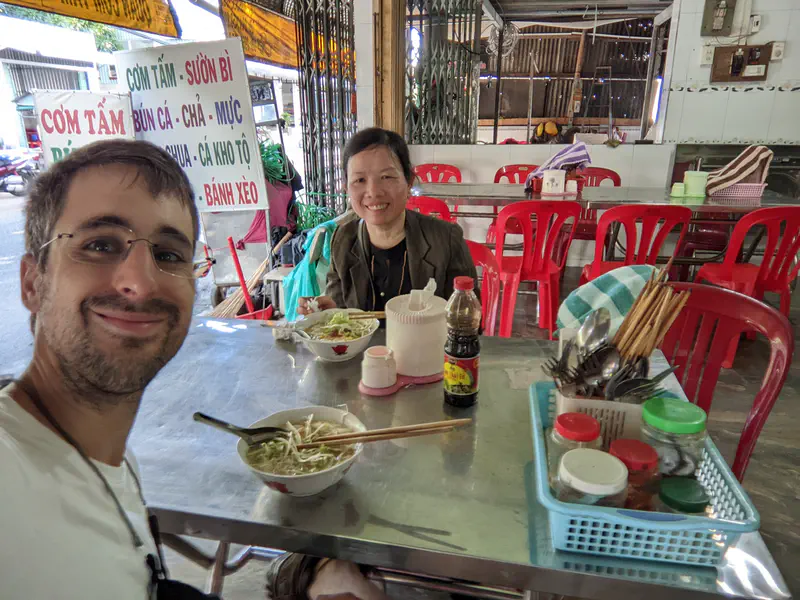 Two people smiling at a metal table with bowls of noodle soup in a casual eatery with red chairs.