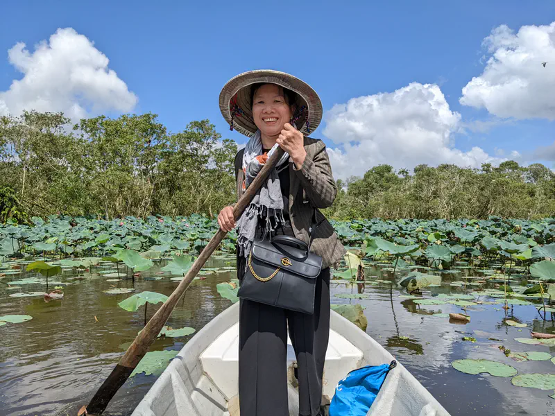 Woman in a conical hat smiling while rowing a boat through a lotus pond under a blue sky.