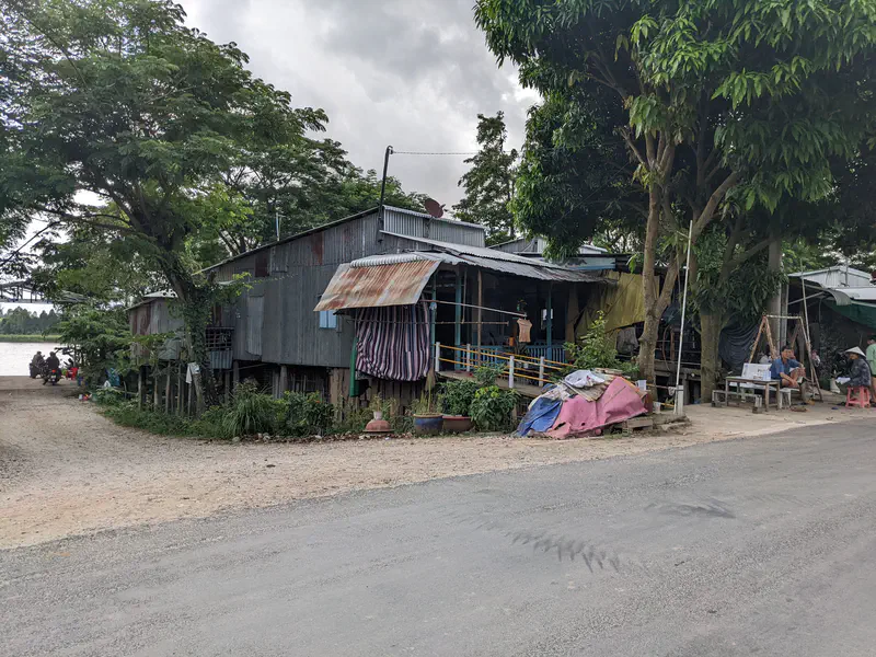 Rustic tin-roofed house on stilts beside a road with trees and people sitting outside under cloudy skies.